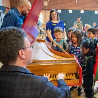 A man plays a red harpsichord for a few dozen elementary-aged school children